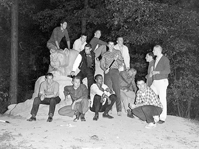 A group of 12 male students sit on and around the Nittany Lion Shrine at night.