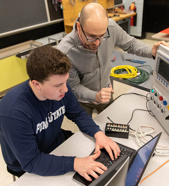 Penn State New Kensington faculty member Joe Cuiffi looks over a student's shoulder at a laptop screen in a robotics lab.