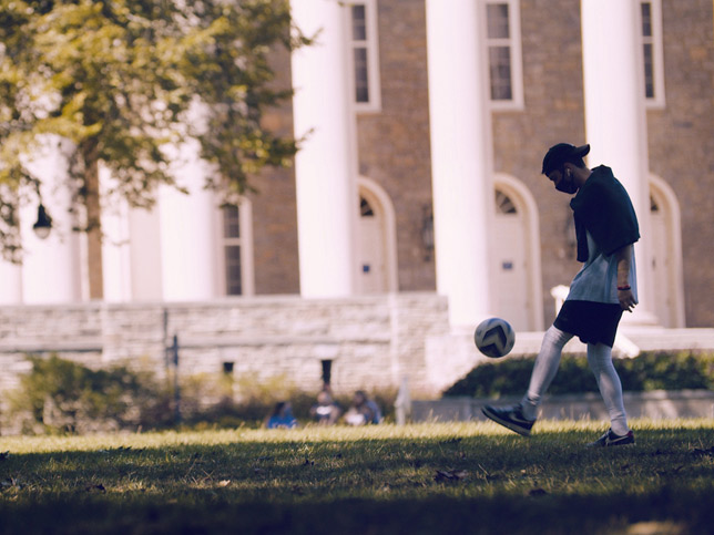 masked student dribbling soccer ball in front of old main.