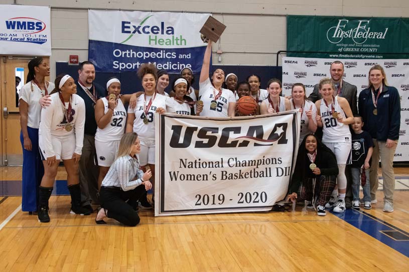 The Beaver Women’s Basketball team, coaches, and staff pose with a banner that says USCAA National Champions Women’s Basketball D2 2019-2020. One student holds a trophy aloft.
