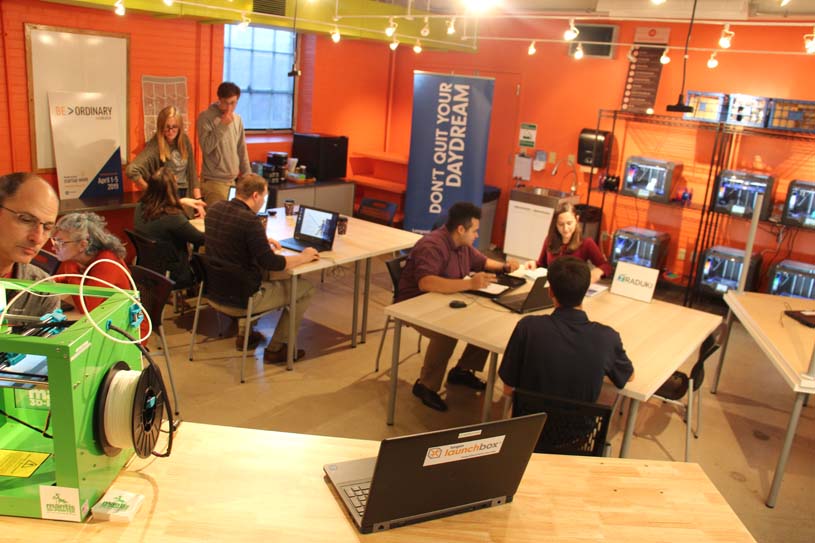 Students seated at tables conversing and working with one in the foreground at a 3D-printer in the Penn State Berks LaunchBox.