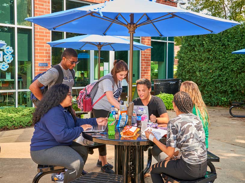 A group of students work on separate devices as they sit at a table under an umbrella outside of the Student Community Center on Penn State Greater Allegheny’s campus.