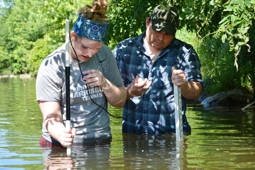 Two students wading in water with measuring instruments while doing research in Conoy Creek near the Penn State Harrisburg campus.