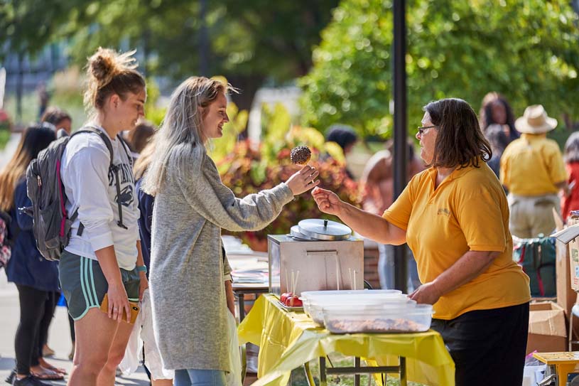 Students stand at an event table as a staff member hands one a candy apple at Fall Fest on the Penn State Harrisburg campus.