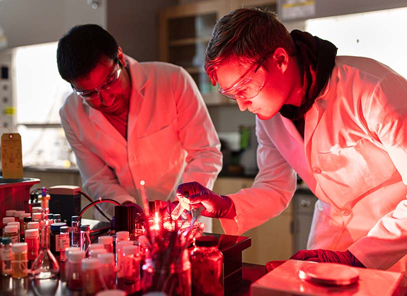 A Penn State Hazleton student and faculty member leaning in towards laboratory equipment that is emitting a red glow.
