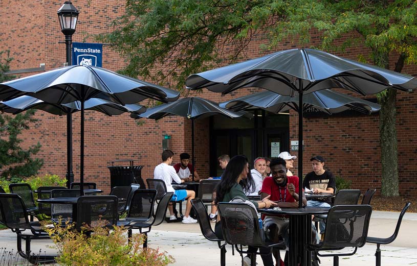 Students hanging out at tables with umbrellas outside Physical Education Building on Hazleton’s campus.