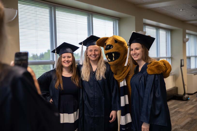 Three New Kensington students wearing commencement regalia stand with Penn State's Nittany Lion mascot as another student takes a photo.