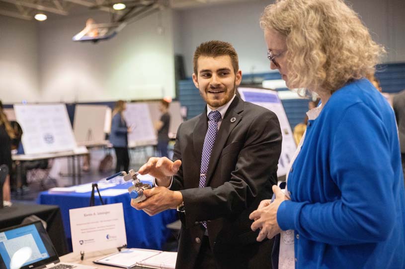 Student in a suit holds a metal part from a research project while talking to a faculty member during a research fair at Penn State New Kensington.