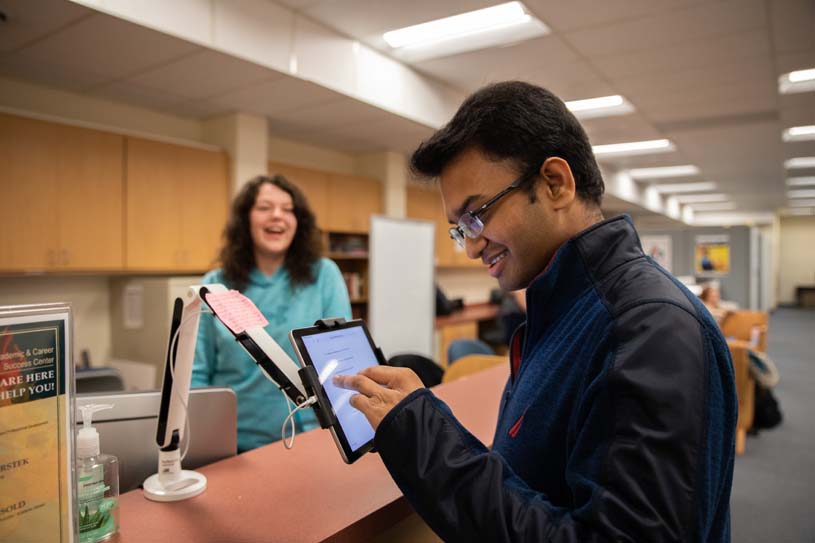 A student stands behind a desk smiling as another students signs into the New Kensington Student Success Center using a touch screen.