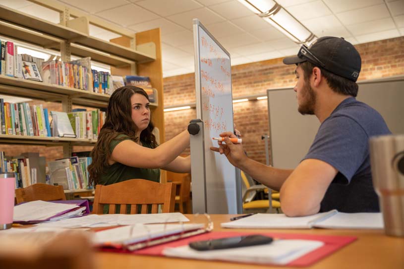 Two students sit at a table in the library at Penn State New Kensington and write math equations on dry erase board.