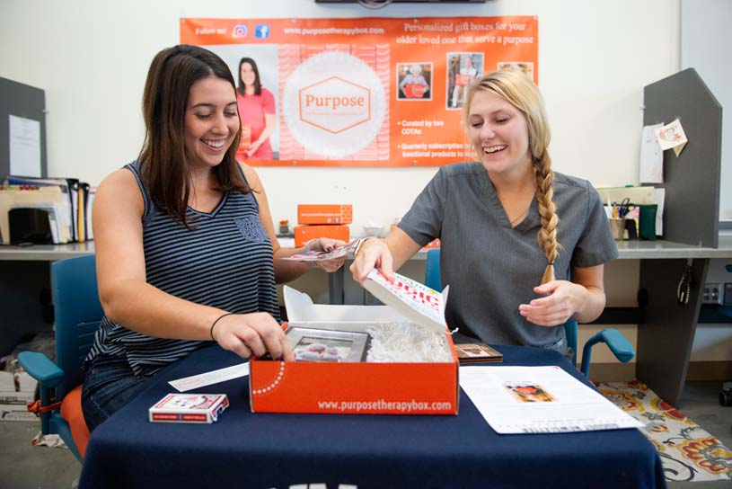 Shenango students and pack an orange box to prepare for shipping. In the background, a banner advertises their business Purpose Therapy Box, LLC.