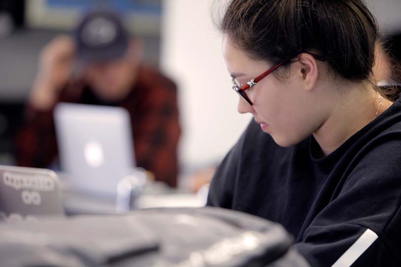 In the foreground, a student looks down at her laptop in class with other students