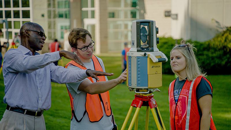 Two students in neon vests using a survey engineering 360 camera with a faculty adviser.