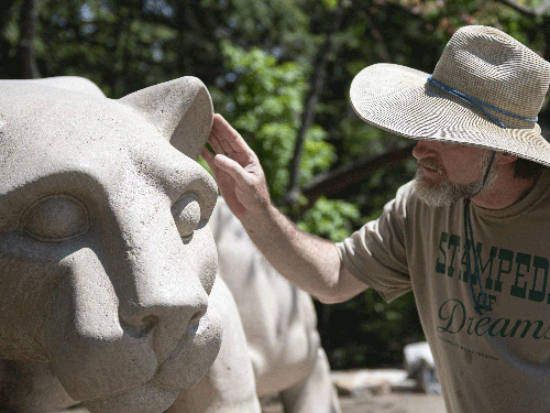 A photo of a sculptor, donned in a straw hat, gingerly reaching out his hand to touch the left ear of the Lion Shrine.