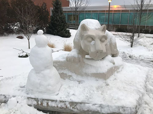 A snowman sits in front of a snow-covered Nittany Lion Shrine, with more snow on the ground, evergreens in the background, and a school building.