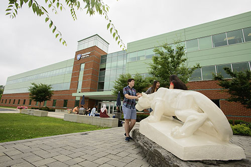 In the right corner, the Nittany Lion shrine faces away from the camera and towards the left. Students pat the front of the statue, and there is a Penn State Lehigh Valley building visible in the background.