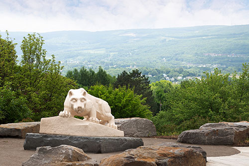 There are boulders in the foreground, the Nittany Lion Shrine in the mid-ground, and lush trees, blue skies, and mountains in the background.