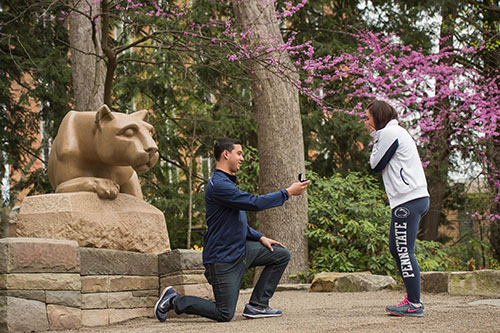 On the left, there is the side profile of the Nittany Lion Shrine. A man, on his knee, is holding out a ring box to a woman who grasps her face in emotion. In the background are the green and pink flowering trees of spring.