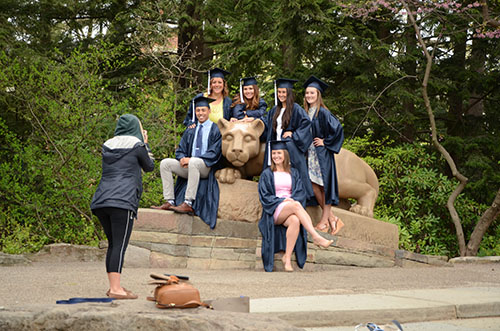 A wide photo of the Nittany Lion Shrine area. There is a woman in the foreground taking a photo of six students sitting around the Lion in their navy caps and gowns for graduation.