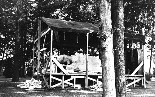A black and white photo of two in-progress lion shrines sitting up on a covered wooden platform.