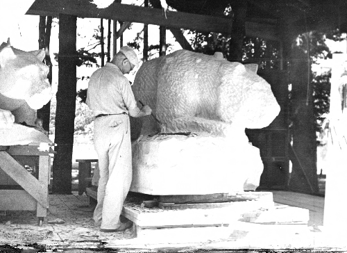 Joseph Garratti chisels out a rough shape of the Lion Shrine. There is a second lion shrine behind him, and we see only the head. The one he is working on is less defined.