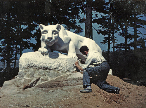 A color photo of Heinz Warneke kneeling in the dirt, working on the Nittany lion shrine sculpture. The shrine is facing the camera.