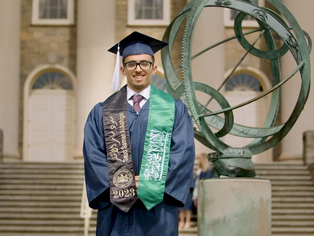A student in graduation regalia in front of Old Maine at Penn State