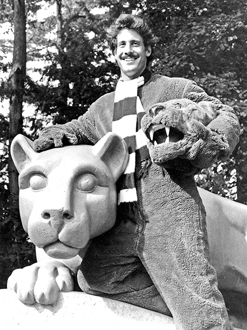 Nittany Lion Mascot Norm Constantine leans up against the Lion Shrine in his mascot costume while holding the headpiece in his arm.