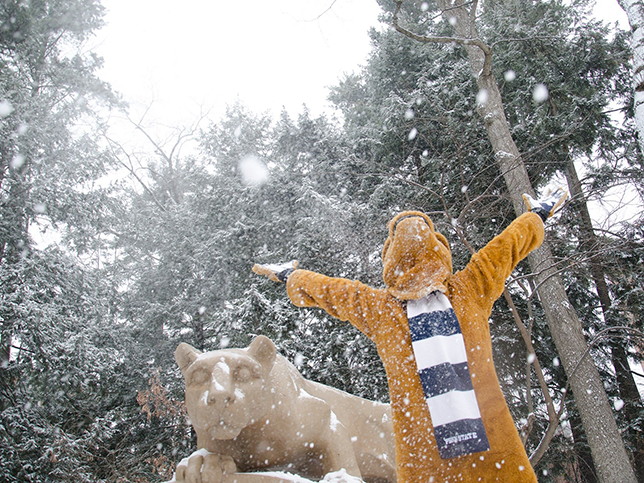 The Nittany Lion mascot stands with its arms stretched wide, throwing snow. Behind it, you can see the lion shrine and evergreen trees covered in snow.