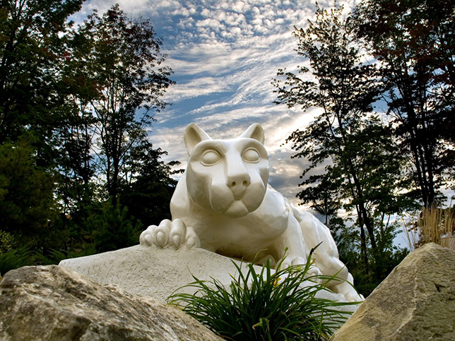 Some rocks pop through the foreground with green plants, the Nittany Lion Shrine looks over those rocks and down at the camera. Behind the lion, there is a blue sky and clouds and lush trees.