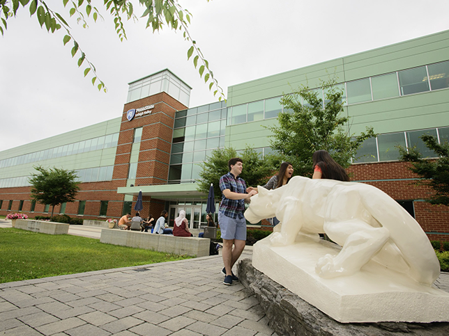 In the right corner, the Nittany Lion shrine faces away from the camera and towards the left. Students pat the front of the statue, and there is a Penn State Lehigh Valley building visible in the background.