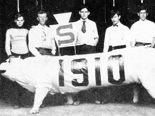 A black-and-white photo of the paper maché lion with 1910 written on the side. There are six male students standing behind it, and a large sign with an 'S' on it.