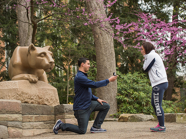 On the left, there is the side profile of the Nittany Lion Shrine. A man, on his knee, is holding out a ring box to a woman who grasps her face in emotion. In the background are the green and pink flowering trees of spring.