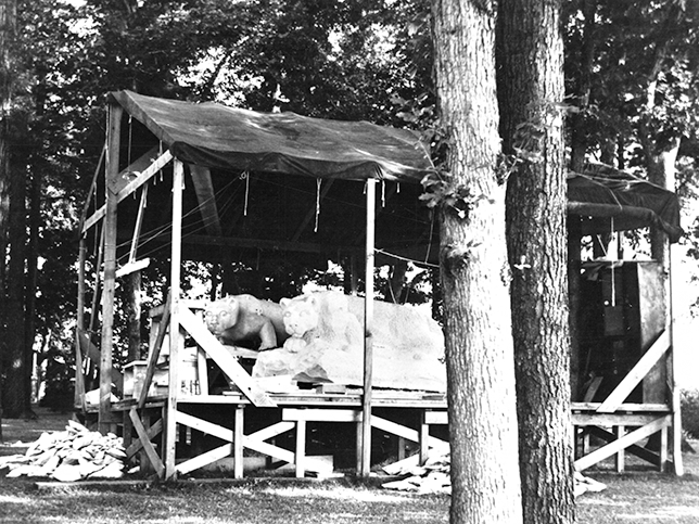 A black and white photo of two in-progress lion shrines sitting up on a covered wooden platform.