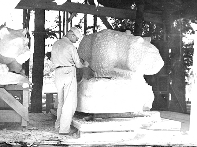 Joseph Garratti chisels out a rough shape of the Lion Shrine. There is a second lion shrine behind him, and we see only the head. The one he is working on is less defined.