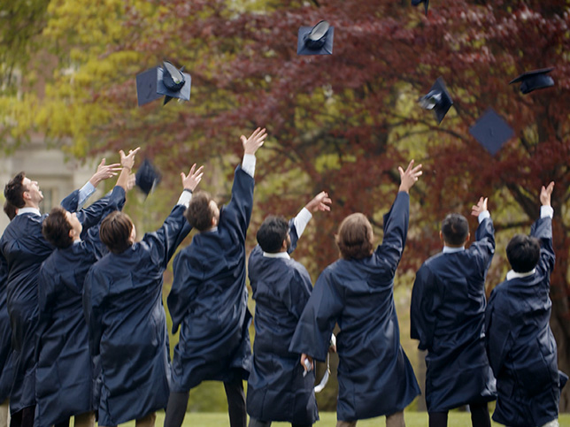 Eight graduates wearing regalia and throwing their caps into the air