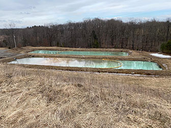 A rectangular blue pond in a brown winter field with a wall of bare trees behind.