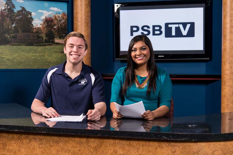 A male and a female student sitting behind a PSB TV news desk